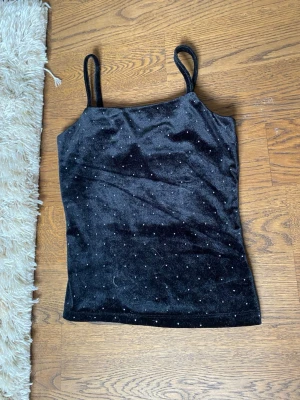 Svart glittrig linnetopp - Snygg svart linnetopp med tunna axelband och glittriga detaljer. Toppen har en mjuk, sammetsliknande yta och är perfekt för att lyfta vilken outfit som helst. Passar dig som gillar att sticka ut lite extra.