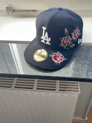 Svart LA New Era keps med rosbrodyr - Säljer en svart New Era 59FIFTY keps med LA-logga i vitt framtill och broderade röda rosor med gröna blad på sidan och skärmen. Klassisk flat brim och sticker kvar på skärmen. Perfekt för dig som vill sticka ut med snygga detaljer. Custom handgjord 