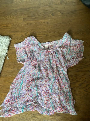 Blommig kortärmad blus i chiffong - Söt blus i tunn chiffong med blommigt mönster i rosa, lila, rött och turkost. Modellen har korta, fladdriga ärmar och rund halsringning med liten rosett. Perfekt för en färgglad och luftig look.