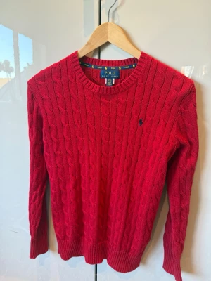 Röd kabelstickad tröja Polo Ralph Lauren - Röd kabelstickad tröja från Polo Ralph Lauren med rund halsringning och diskret blå logga broderad på bröstet. Tröjan har ribbade muddar vid ärmslut och nederkant. Klassisk och tidlös design i mjukt material.