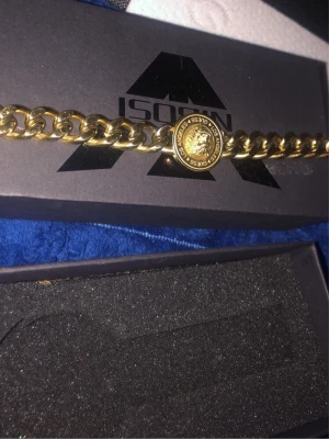 Guldfärgat halsband från Guess - Snyggt guldfärgat halsband från Guess med grov kedja och ett runt hänge med lejonhuvud och texten 'Guess Los Angeles'. Perfekt accessoar för att lyfta din outfit med en lyxig touch. Kommer i original ask.