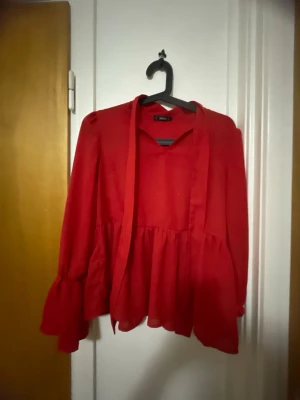 Röd volangblus från Shein - Trendig röd blus från Shein med volang nedtill och långa, vida ärmar. Blusen har en lös passform och knytband vid halsen för extra detalj. Perfekt för dig som vill sticka ut med färg och stil.