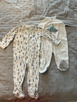 Två vita baby-pyjamasar med mönster - Säljer två söta baby-pyjamasar i vitt. Ena har svarta träd- och husmönster samt tryckknappar framtill, den andra har grå-randiga ärmar, fötter och ett blått tryck med luftballong. Båda har långa ärmar och täckta fötter, perfekta för mysiga nätter.