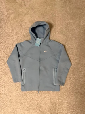 Ljusblå Nike NOCTA hoodie - Snygg ljusblå hoodie från Nike NOCTA-kollektionen med dragkedja framtill och två fickor med dragkedja. Hoodien har en minimalistisk design med vit Nike-logga på bröstet och justerbar huva. Perfekt för en chill och sportig look. Storlek S men sitter lite tight. Priset kan diskuteras, och fler bilder kan skickas 👍