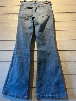 Ljusblå utsvängda jeans - Säljer ett par ljusblå jeans med utsvängda ben och klassisk femficksdesign. Jeansen är tillverkade i mjukt denimtyg. Perfekta för dig som gillar retrostil och vill ha ett par riktigt snygga jeans med 70-talsvibbar. Strl 26/32