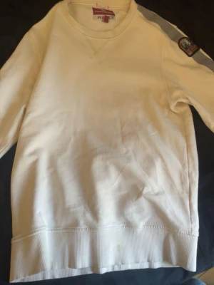 Vit sweatshirt från Parajumpers - Vit sweatshirt från Parajumpers med rund halsringning och ribbade muddar. Tröjan har en grå diagonal rand över axeln och ett broderat Parajumpers-märke på ena ärmen. Tillverkad i mjukt bomullsmaterial för en avslappnad look. S funkar också men då kommer den sitta tight