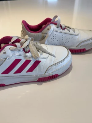 Vita och rosa Adidas sneakers - Snygga vita sneakers från Adidas med rosa detaljer och klassiska tre ränder på sidan. Skorna har vita snören, rosa insida och sula samt perforerade partier på sidan för ventilation. Tillverkade i syntetmaterial och textil för en sportig look.