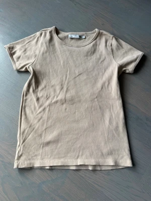 Beige ribbad topp från 157 - Säljer en beige ribbad topp från 157 med korta ärmar och rund hals. Toppen är i ett mjukt och stretchigt material som sitter snyggt på kroppen. Perfekt att styla med jeans eller kjol för en clean look.