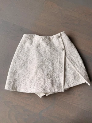 Vita strukturerade skorts med knappar - Säljer ett par vita skorts med snygg struktur och omlott-design framtill. Skortsen har två dekorativa silverknappar och resår i midjan bak för extra komfort. Materialet är tjockt och har ett elegant mönster. Perfekt för dig som vill ha kjol-look men shorts-känsla.