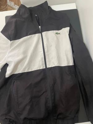 Svartvit vindjacka från Lacoste med byxor - Snygg svart och vit vindjacka från Lacoste med dragkedja framtill och hög krage. Jackan har det klassiska gröna krokodilmärket på bröstet och en stor krokodil på ena ärmen. Tillverkad i lätt och tåligt material, perfekt för blåsiga dagar.