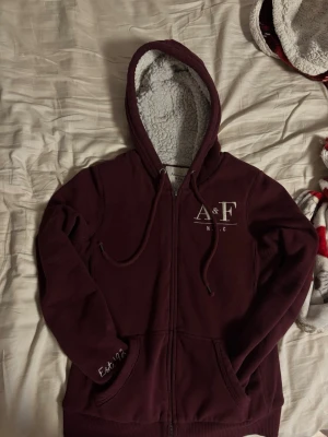 Vinröd zip hoodie Abercrombie & Fitch - Mysig vinröd hoodie från Abercrombie & Fitch med dragkedja och vit logga på bröstet. Fodrad med fluffigt vitt teddyfoder i huvan och insidan, samt snören i huvan. Perfekt för chill dagar och har fickor framtill.