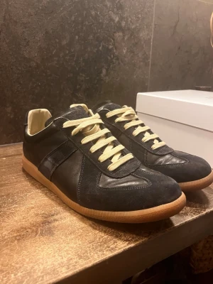 Marinblå Maison margielas  - Säljer ett par marinblå sneakers från Maison Margiela med beige gummisula och kontrasterande beigea skosnören. Skorna har en mix av mocka och läder på ovandelen samt en klassisk låg siluett. De ska vara marinblå men ser lite svarta ut. Endast använda några enstaka gånger. Skriv vid frågor eller funderingar!