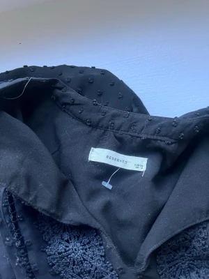 Svart blus med struktur från Reserved - Snygg svart blus från Reserved med coola virkade detaljer framtill och små prickar i tyget. Blusen har krage, långa ärmar och resår i midjan för en skön passform. Perfekt för dig som gillar unika detaljer och stilren look.