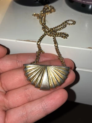 Guldigt halsband från Karl lagerfeld - Unikt halsband i guldfärgad metall med ett stort solfjäderformat hänge. Kedjan är grov och matchar hängets retrostil. Perfekt för dig som gillar statement-smycken och vill sticka ut med en cool accessoar från Karl Lagerfeld 