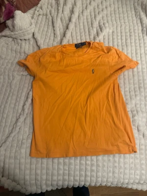 Orange t-shirt från Polo Ralph Lauren - Snygg orange t-shirt från Polo Ralph Lauren med klassisk passform och liten broderad logga på bröstet. Tillverkad i mjuk bomull och har rund hals samt korta ärmar. Perfekt för en avslappnad och stilren look.