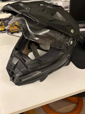 Svart crosshjälm med visir - Säljer en svart crosshjälm från DXR med integrerat klart visir och skärm. Hjälmen har en modern, kantig design och är tillverkad i slitstark plast med ventiler och vadderad insida för extra komfort. Några repor här och var men inte mycket.