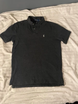 Ralph lauren piké - Klassisk mörkgrå pikétröja från Polo Ralph Lauren i modellen Classic Fit. Tröjan har korta ärmar, krage med två knappar och den ikoniska broderade loggan på bröstet. Tillverkad i mjuk bomull för en bekväm känsla.