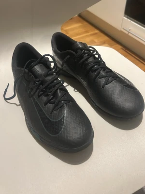 Svarta Nike fotbollsskor med snörning - Säljer ett par helsvarta fotbollsskor från Nike med klassisk låg profil och snörning. Skorna har en diskret Nike-logga på sidan och ovandelen är tillverkad i syntetmaterial med mönstrad yta för bättre bollkontroll. Perfekta för dig som vill ha stilrena och funktionella skor på planen.
