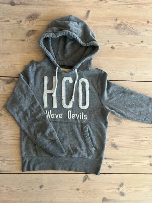 Hollister Hoodie - Grå hoodie från Hollister med stor vit text 'HCO Wave Devils' på bröstet. Klassisk ficka framtill och justerbar huva med snören. Tillverkad i mjukt material som passar perfekt för en avslappnad stil. Storlek S!🙌🙌
