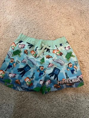 Minecraft badbyxor med figurmönster - Säljer ett par ljusblå badbyxor med elastisk midja och snöre, täckta av färgglada Minecraft-figurer som Steve, Alex, bläckfiskar, sköldpaddor och delfiner. Tryckt Minecraft-logga framtill. Perfekta för dig som älskar gaming och vill sticka ut på stranden. Det behövs dock sys en raksöm mellan benen där de börja gå sönder, men annars i väldigt gott skick!