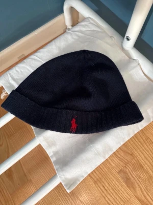Mörkblå mössa från Polo Ralph Lauren - Snygg mörkblå mössa från Polo Ralph Lauren i mjuk och tvättbar merinoull. Klassisk ribbad kant och ikoniska röda logotypen broderad framtill. Perfekt accessoar för kalla dagar och enkel att matcha med olika outfits.