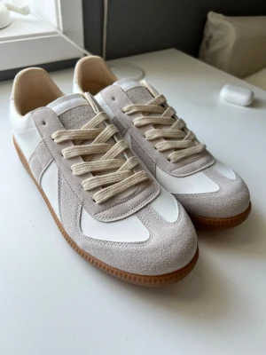 Vita och beige sneakers Maison Margiela - Stilrena sneakers från Maison Margiela i vitt och beige med ovandel i mocka och läder. Klassisk gummisula i brunt och snygga detaljer på sidan. Skorna har låga snitt och beigea skosnören som matchar designen perfekt. Använda en gång
