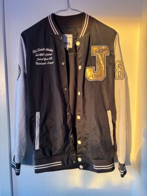 Svart och vit bomberjacka Jeel DNM - Snygg svart och vit bomberjacka från Jeel DNM med broderade detaljer, stora bokstäver och siffror på bröst och ärm. Jackan har ribbade muddar, tryckknappar och kontrastfärgade ärmar. Perfekt för en sportig och trendig look.