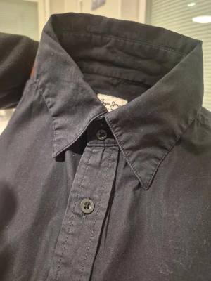 Pepe Jeans M23140 New Eric 099svart skjorta, storlek L - Klassisk svart skjorta från Pepe Jeans med lång ärm och knappar framtill. Skjortan har en stilren design med normal passform och är tillverkad i mjuk bomull. Perfekt till jeans eller chinos för en clean look.