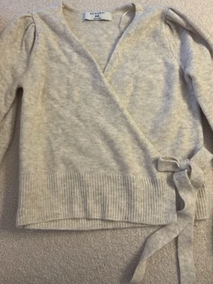 Ljusgrå omlottkofta från Primark - Supersnygg ljusgrå stickad omlottkofta från Primark med långa ärmar och knytband i sidan. Koftan har ribbstickade muddar vid ärmslut och nederkant, samt lätt puff vid axlarna för en trendig look. Perfekt att slänga över en topp för extra stilpoäng.