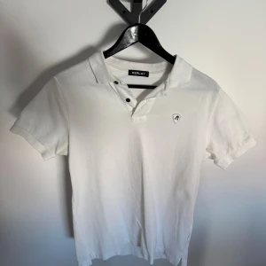 Vit pikétröja från Replay - Klassisk vit pikétröja från Replay med korta ärmar och krage. Tröjan har knappar vid halsen och en liten logga på bröstet. Perfekt till en avslappnad men ändå stilren look.