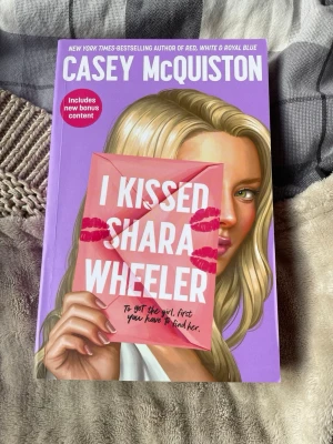I Kissed Shara Wheeler bok - En färgstark pocketbok med lila omslag och illustration av en blond tjej som håller ett brev. Titeln är 'I Kissed Shara Wheeler' och boken är skriven av Casey McQuiston. Omslaget har kyssmärken och boken innehåller bonusmaterial.