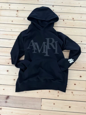 Svart hoodie från Amiri med tryck - Snygg svart hoodie från Amiri med stor logga i svart på bröstet och vit detalj på ena ärmen. Klassisk känguruficka framtill och stor huva. Perfekt för dig som gillar streetwear och vill ha något stilrent men ändå med edge.