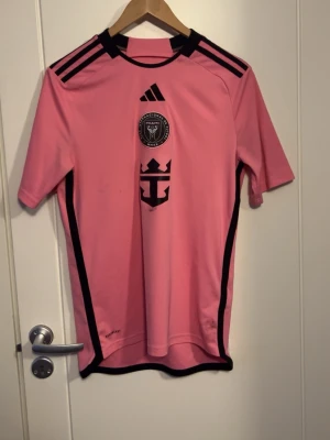 Inter Miami Messi matchtröja rosa - Säljer en rosa Inter Miami matchtröja från Adidas med svarta detaljer. Tröjan har klubbmärke och sponsorlogga på bröstet samt Messi och nummer 10 på ryggen. Tillverkad i lätt och ventilerande material, perfekt för fotboll eller träning.