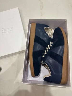 Marinblå sneakers Maison Margiela - Snygga marinblå sneakers från Maison Margiela med ovandel i mocka och läder, vita skosnören och klassisk gummisula i brunt. Skorna har en stilren och tidlös design som passar perfekt till streetwear och casual outfits.