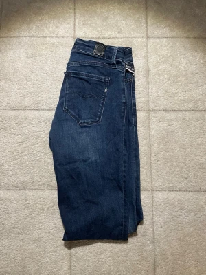 Mörkblå slim jeans från Replay - Säljer ett par mörkblå slim jeans från Replay med klassisk femficksdesign och dragkedja. Jeansen har en snygg tvättad look och smal passform som sitter tajt längs benen. Perfekta för en trendig och avslappnad stil.