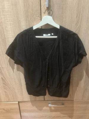 Svart kortärmad blus med knappar - Svart blus med v-ringning och korta ärmar. Modellen är lite croppad och har knappar framtill. Tillverkad i ett lätt, något glansigt material som ger en avslappnad men ändå stilren look. Perfekt att matcha med både jeans och kjol.