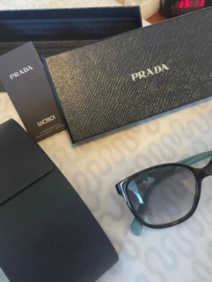 Svarta solglasögon från Prada - Snygga svarta solglasögon från Prada med klassisk cat-eye form och mörka glas. Bågarna har en elegant svart finish med diskret Prada-logga på sidan. Kommer med originalfodral och ask, perfekt för dig som vill ha en lyxig accessoar.