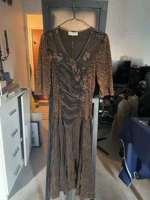 Brun långklänning från Delvalle - Unik brun långklänning från Delvalle med v-ringning, trekvartsärm och dekorativa blommiga broderier. Klänningen har draperad front och volangdetaljer nedtill för en bohemisk look. Perfekt för dig som vill sticka ut med en kreativ och konstnärlig stil.