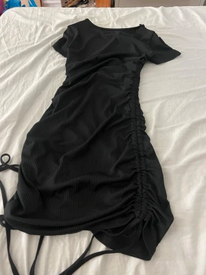 Svart ribbad kortklänning med snörning - Svart kortklänning med ribbad struktur och snörning längs sidan som ger en snygg rynkeffekt. Klänningen har korta ärmar och en enkel rund halsringning. Perfekt för dig som gillar en stilren och trendig look.
