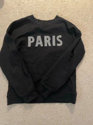 Svart sweatshirt PARIS från MNG - Svart sweatshirt från MNG med glittrig text 'PARIS' framtill. Klassisk rund halsringning och ribbade muddar vid ärmslut och nederkant. Tillverkad i mjukt material som är skönt. Perfekt för dig som gillar en stilren look.