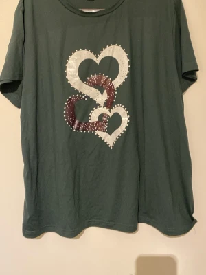 Grön t-shirt med hjärtmotiv - Mörkgrön t-shirt med korta ärmar och tryck av tre stora hjärtan i vitt och vinrött på framsidan. Hjärtana är dekorerade med små vita prickar runtom. Skön passform och mjukt material, perfekt till vardags.