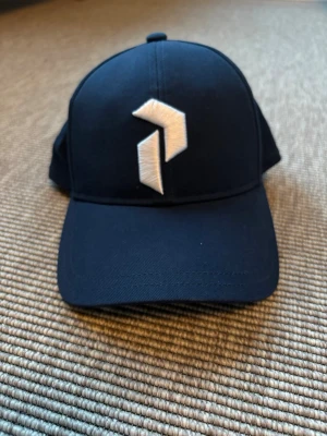 Peak Performance- Mörkblå juniorkeps  - Snygg mörkblå juniorkeps från Peak Performance med vit broderad logga framtill. Klassisk böjd skärm och justerbar passform. Tillverkad i slitstarkt material som passar perfekt till streetwear eller sportiga outfits.