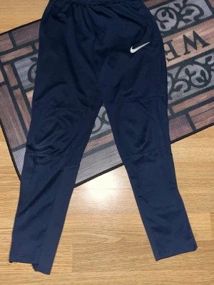 Mörkblå Nike träningsbyxor - Säljer ett par mörkblå träningsbyxor från Nike med vit logga på framsidan. Byxorna har elastisk midja och är tillverkade i ett lätt och stretchigt material som passar perfekt för sport och träning. De har en smal passform och paneler över knäna för extra komfort.