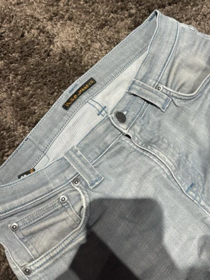 Ljusblå jeans från Nudie Jeans - Ljusblå jeans från Nudie Jeans med klassisk femficksdesign och silverfärgade nitar. Jeansen har en rak passform och är tillverkade i mjukt denim-material. Perfekta för dig som gillar en avslappnad och stilren look.