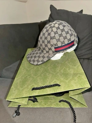 Gucci beige keps med GG-mönster - Säljer en beige keps från Gucci med det klassiska GG-monogrammet över hela kepsen. På sidan finns en ikonisk röd och marinblå rand. Kepsen har böjd skärm och är tillverkad i canvasmaterial. Perfekt för dig som vill ha en lyxig och trendig accessoar.