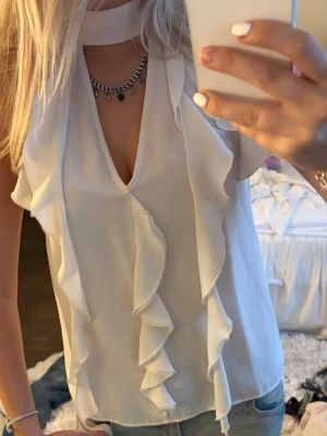 Vit volangblus med v-ringning - Säljer en snygg vit blus med v-ringning och volangdetaljer framtill. Blusen är ärmlös och har en dekorativ halsringning som ger en trendig look. Perfekt att styla med jeans eller kjol för en chic outfit.