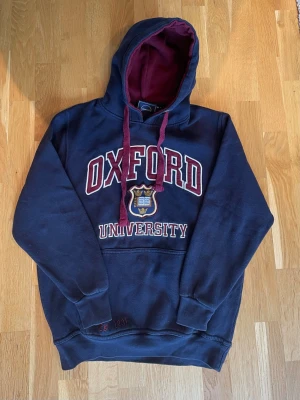 Vintage Oxford University hoodie - Mörkblå hoodie från Oxford University med stor logga och text framtill. Hoodien har vinröda snören och insida av huvan i matchande vinröd färg. Klassisk känguruficka och mjukt fleece-material gör den extra bekväm. Perfekt för en avslappnad och stilren look.