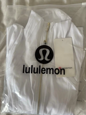 Vit defyne jacket med guld zip up från Lululemon - Snygg vit defyne jacket från Lululemon med guldfärgad dragkedja och logga på bröstet. Tillverkad i ett stretchigt och lätt material med inslag av Lycra för extra komfort och rörelsefrihet. Perfekt för träning eller som ett sportigt lager.