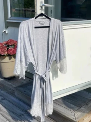 Ljusgrå morgonrock med spetsdetaljer - Snygg ljusgrå morgonrock med knytband i midjan och breda ärmar. Spetsdetaljer vid ärmslut och nederkant ger en romantisk touch. Perfekt för mysiga hemmakvällar eller som loungewear. Mjuk och lätt i materialet.