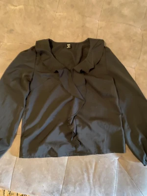 Svart blus med volang från Emery Rose - Stilren svart blus från Emery Rose i storlek XS. Blusen har långa ärmar och en snygg volangdetalj längs halsringningen och framtill. Tillverkad i ett lätt tyg som ger ett elegant intryck. Perfekt för dig som gillar trendiga detaljer och klassisk svart.